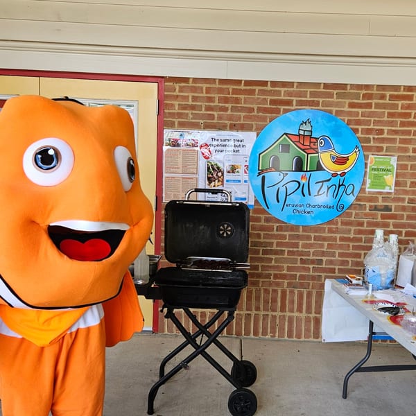 An orange mascot stands next to a grill and a Pipilinka sign at an outdoor event.
