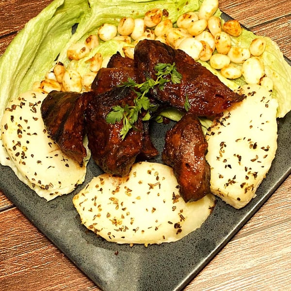 Peruvian dish with dark glazed meat, white potatoes, corn, and lettuce on a dark plate.